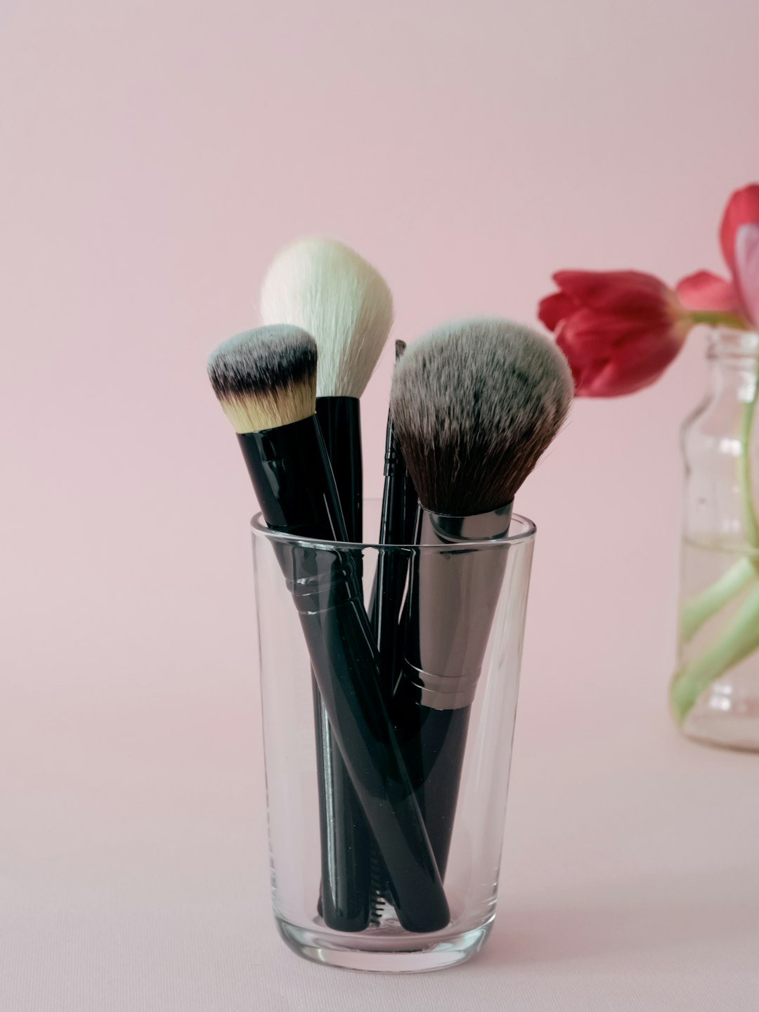 Set of makeup brushes on a pink background by Makeup Brands a glass filled with makeup brushes next to a vase with tulips